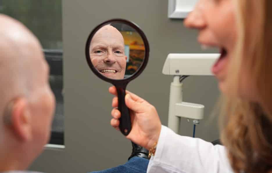 Smiling All-on-X patient Dennis looking at his reflection in a handheld mirror during a follow-up visit at Longmont Dental Loft.