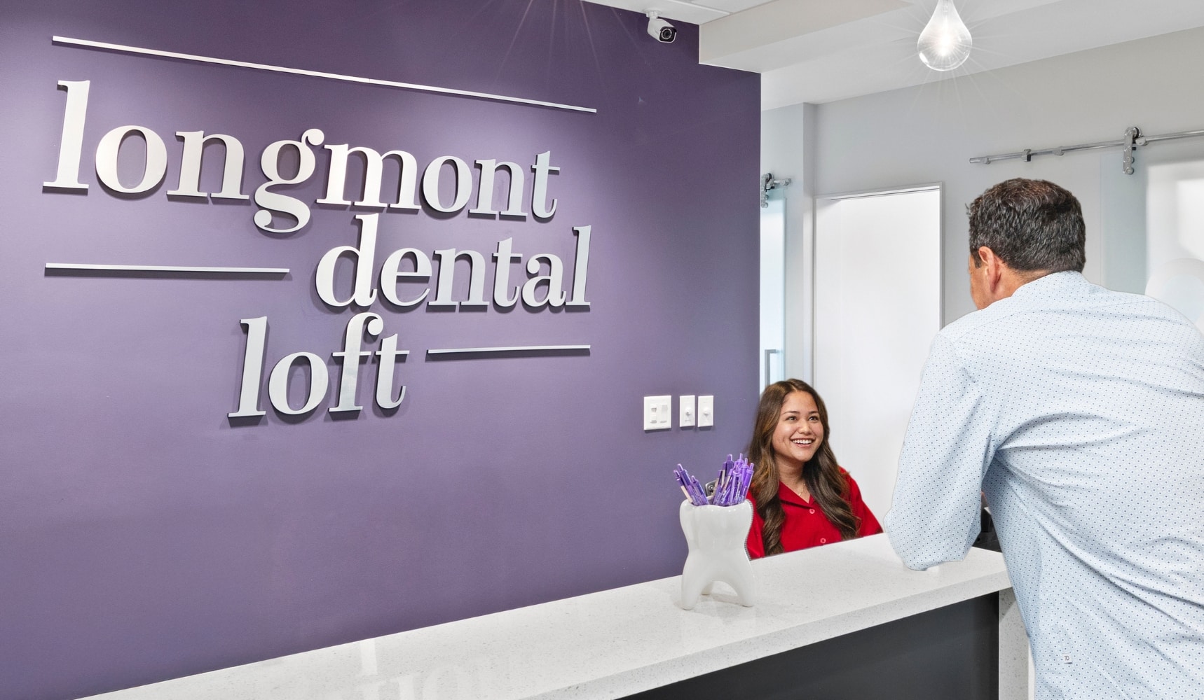 Patient coordinator smiling while speaking with a patient at the Longmont Dental Loft front desk, with the office logo on a purple wall behind them.